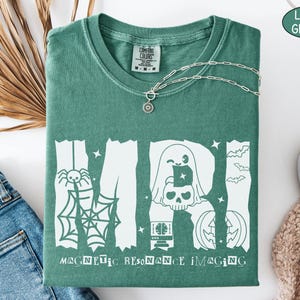 May include: A light green short-sleeved t-shirt featuring a white graphic print of the letters MRI. The design includes a spider, web, ghost, skull, computer monitor, and pumpkin. The text Magnetic Resonance Imaging is printed below the letters.