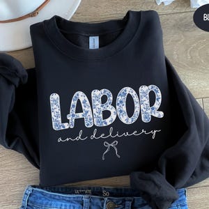 Op de afbeelding: Zwarte sweatshirt met de woorden "LABOR and delivery" in witte letters en bloemenletters. Een witte strik staat onder de tekst. De sweatshirt is opgevouwen op een blauwe jeans.