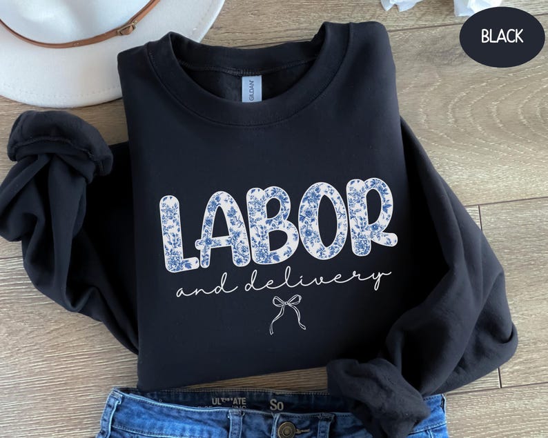 Op de afbeelding: Zwarte sweatshirt met de woorden "LABOR and delivery" in witte letters en bloemenletters. Een witte strik staat onder de tekst. De sweatshirt is opgevouwen op een blauwe jeans.