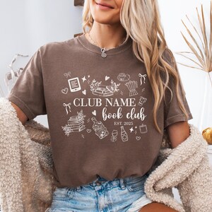 May include: A brown t-shirt with white graphic illustrations and text. The text reads "CLUB NAME book club EST. 2025". The graphic includes books, a coffee pot, wine, and other book-related icons. The shirt is worn by a person.