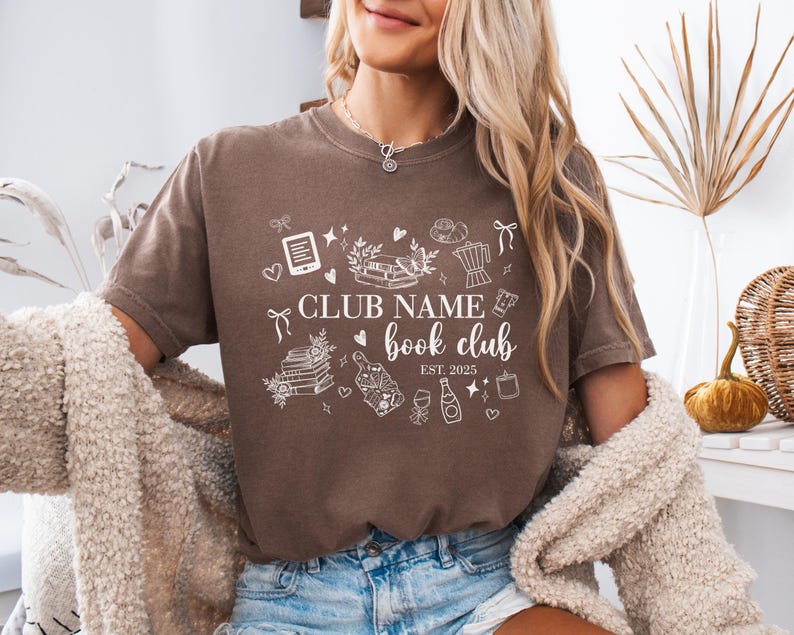 May include: A brown t-shirt with white graphic illustrations and text. The text reads "CLUB NAME book club EST. 2025". The graphic includes books, a coffee pot, wine, and other book-related icons. The shirt is worn by a person.