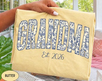 May include: A butter yellow t-shirt featuring the word GRANDMA in large, block-style letters filled with a blue and white floral pattern. Below the word, the text Est. 2026 is printed in a simple, dark blue cursive script font.