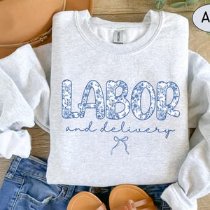 Op de afbeelding: Een lichtgrijs sweatshirt met de tekst "LABOR and delivery" in een blauw bloemenpatroon. De afbeelding toont ook een blauwe jeans, sandalen en een bruine hoed, wat een casual stijl suggereert.