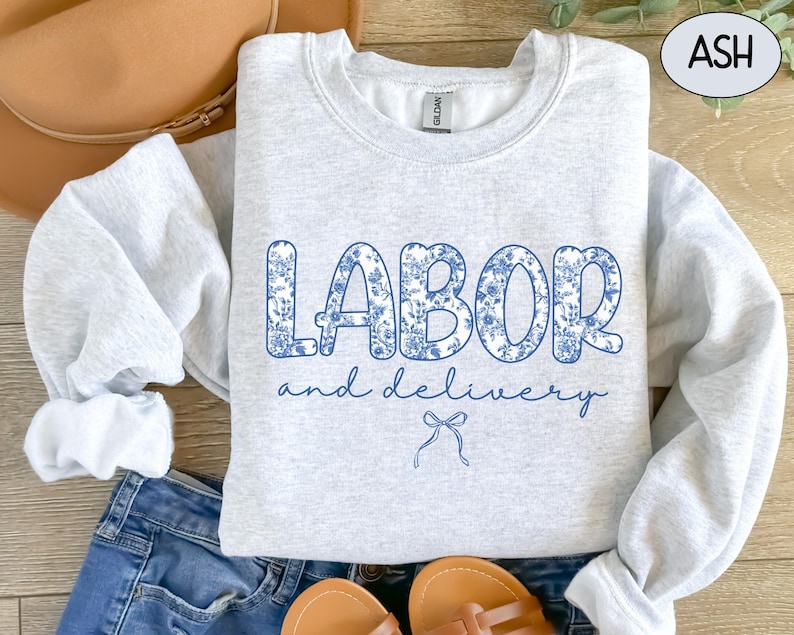 Op de afbeelding: Een lichtgrijs sweatshirt met de tekst "LABOR and delivery" in een blauw bloemenpatroon. De afbeelding toont ook een blauwe jeans, sandalen en een bruine hoed, wat een casual stijl suggereert.