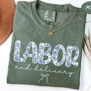 Op de afbeelding: Een mosgroen t-shirt met de woorden "LABOR and delivery" in witte letters en bloemenprint. Een gouden ketting en een bruine hoed zijn ook zichtbaar. Het shirt is gemaakt van een zacht, comfortabel materiaal.