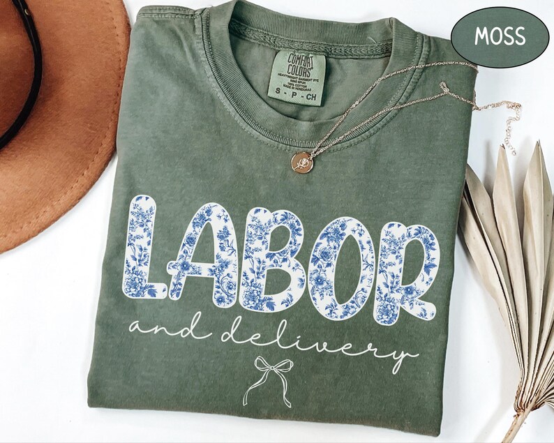 Op de afbeelding: Een mosgroen t-shirt met de woorden "LABOR and delivery" in witte letters en bloemenprint. Een gouden ketting en een bruine hoed zijn ook zichtbaar. Het shirt is gemaakt van een zacht, comfortabel materiaal.