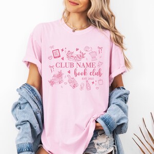 May include: A light pink t-shirt with a pink graphic design featuring the text "CLUB NAME book club EST. 2025". The design includes illustrations of books, hearts, and other book-related items.