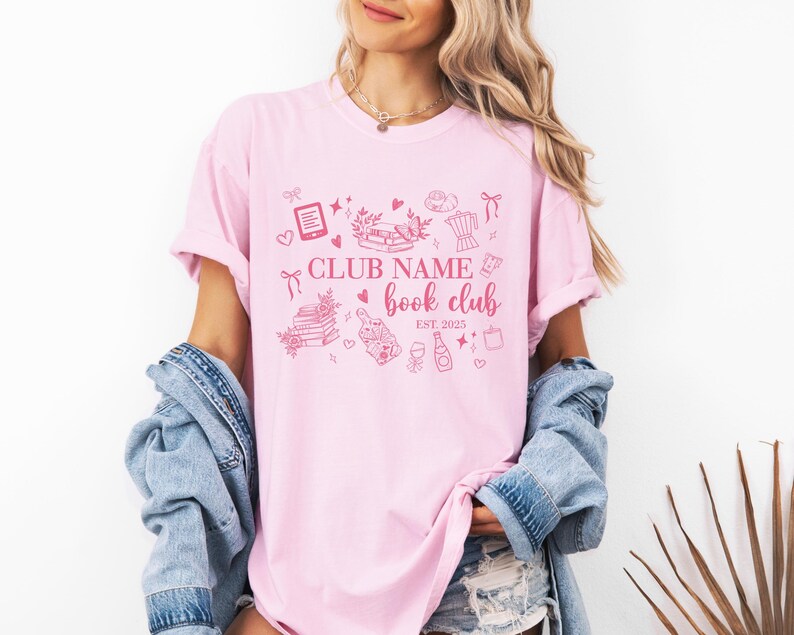 May include: A light pink t-shirt with a pink graphic design featuring the text "CLUB NAME book club EST. 2025". The design includes illustrations of books, hearts, and other book-related items.