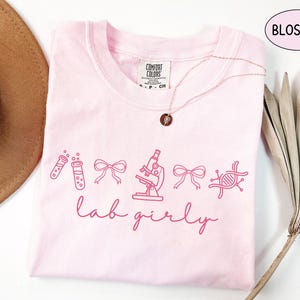 May include: A light pink t-shirt with the word "Blossom" in a pink oval. The shirt features pink line art of lab equipment and the words "lab girly" in cursive. A gold necklace with a pendant is on the shirt.
