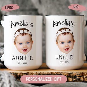 May include: Two white ceramic mugs with black handles and rims. Each mug features a baby's photo, the name "Amelia's", and the words "AUNTIE" and "UNCLE" with "EST. 2025". The mugs are personalized gifts.