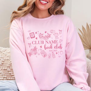 May include: A light pink sweatshirt with a book club design. The design includes the text "CLUB NAME book club EST. 2025" with illustrations of books, hearts, and other book-related items. The sweatshirt is a solid light pink color.