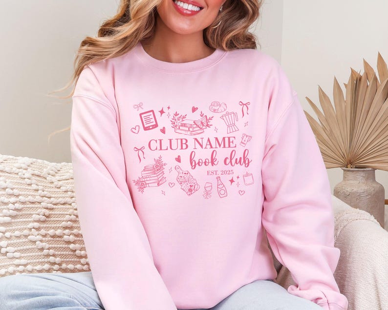 May include: A light pink sweatshirt with a book club design. The design includes the text "CLUB NAME book club EST. 2025" with illustrations of books, hearts, and other book-related items. The sweatshirt is a solid light pink color.