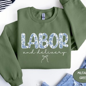 Op de afbeelding: Olijfgroene sweatshirt met de woorden "LABOR" en "and delivery" in witte letters. Het woord "LABOR" is gevuld met een blauw bloemenpatroon. Onder de tekst staat een kleine strikgrafiek. De sweatshirt is op een wit oppervlak gevouwen.