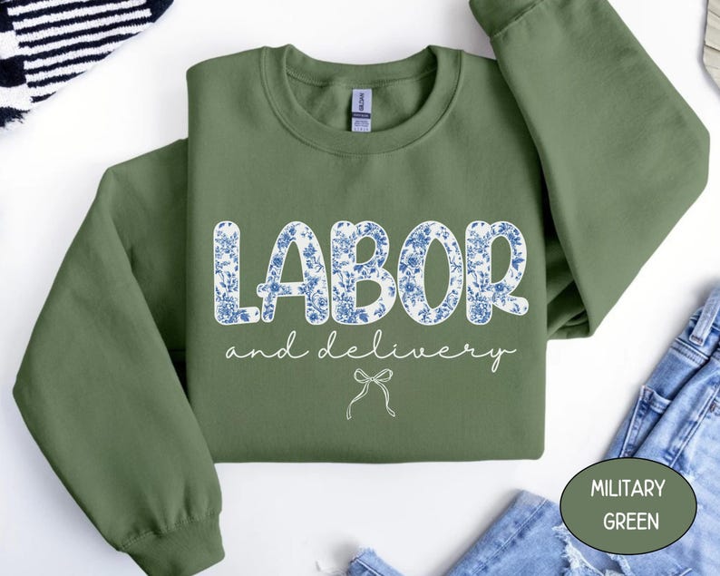 Op de afbeelding: Olijfgroene sweatshirt met de woorden "LABOR" en "and delivery" in witte letters. Het woord "LABOR" is gevuld met een blauw bloemenpatroon. Onder de tekst staat een kleine strikgrafiek. De sweatshirt is op een wit oppervlak gevouwen.