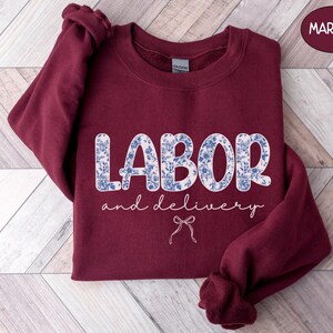 Op de afbeelding: Een bordeauxrode sweatshirt met de tekst "LABOR and delivery" in witte letters en een blauw-witte bloemenprint. Een kleine strik-afbeelding staat onder de tekst. De sweatshirt wordt gepresenteerd op een houten oppervlak.