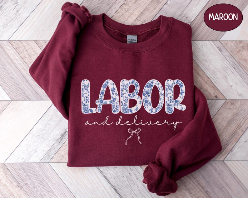 Op de afbeelding: Een bordeauxrode sweatshirt met de tekst "LABOR and delivery" in witte letters en een blauw-witte bloemenprint. Een kleine strik-afbeelding staat onder de tekst. De sweatshirt wordt gepresenteerd op een houten oppervlak.