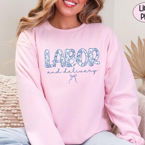 Op de afbeelding: Lichtroze sweatshirt met de tekst "LABOR and delivery" in een blauwe bloemenprint. Een blauwe strik-afbeelding staat onder de tekst. Het sweatshirt is effen lichtroze van kleur.