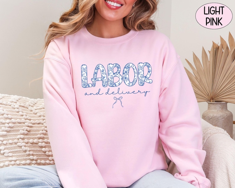 Op de afbeelding: Lichtroze sweatshirt met de tekst "LABOR and delivery" in een blauwe bloemenprint. Een blauwe strik-afbeelding staat onder de tekst. Het sweatshirt is effen lichtroze van kleur.