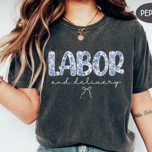 Op de afbeelding: Donkergrijs t-shirt met de woorden "LABOR" en "and delivery" in witte letters. Het woord "LABOR" staat in grote blokletters gevuld met een blauw bloemenpatroon. Een kleine strik-afbeelding staat eronder.