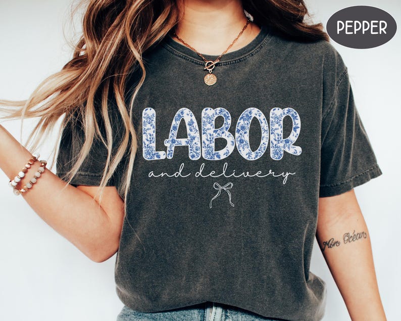 Op de afbeelding: Donkergrijs t-shirt met de woorden "LABOR" en "and delivery" in witte letters. Het woord "LABOR" staat in grote blokletters gevuld met een blauw bloemenpatroon. Een kleine strik-afbeelding staat eronder.