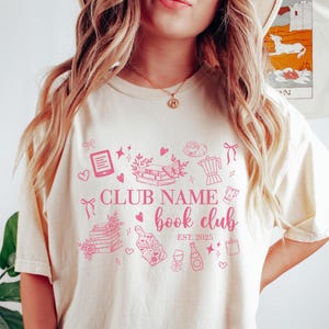 May include: Cream-colored t-shirt with a pink book club design. The design includes illustrations of books, a butterfly, a coffee pot, and the text "CLUB NAME book club EST. 2025".