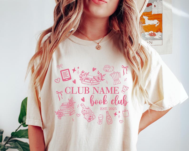 May include: Cream-colored t-shirt with a pink book club design. The design includes illustrations of books, a butterfly, a coffee pot, and the text "CLUB NAME book club EST. 2025".