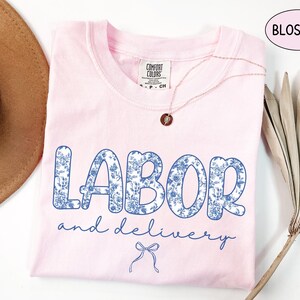 Op de afbeelding: Lichtroze t-shirt met de woorden "LABOR and delivery" in blauwe bloemenprint. Het shirt heeft een ronde hals en korte mouwen. Een gouden ketting met een hanger ligt op het shirt.