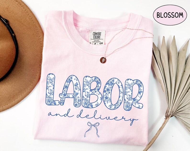 Op de afbeelding: Lichtroze t-shirt met de woorden "LABOR and delivery" in blauwe bloemenprint. Het shirt heeft een ronde hals en korte mouwen. Een gouden ketting met een hanger ligt op het shirt.