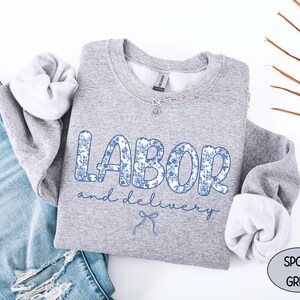 Op de afbeelding: Een grijze sweatshirt met de woorden "LABOR and delivery" in blauwe bloemenprint. De sweatshirt wordt gecombineerd met een blauwe jeans. Een zilveren ketting is over de sweatshirt gedrapeerd.