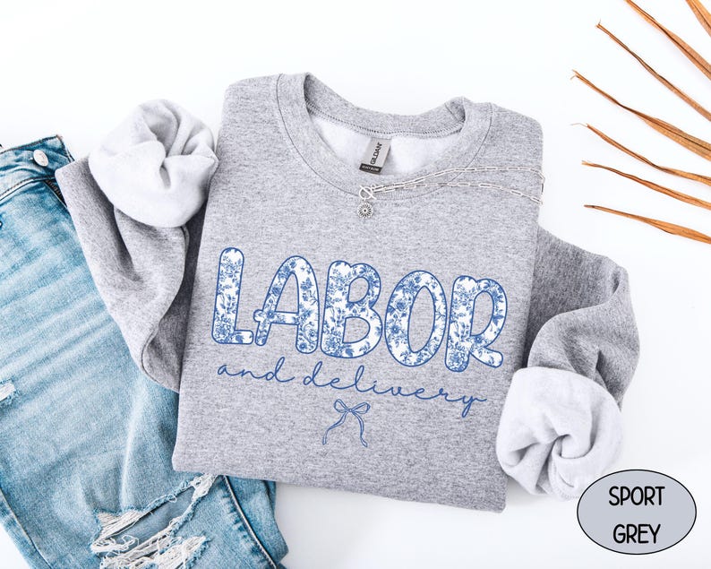 Op de afbeelding: Een grijze sweatshirt met de woorden "LABOR and delivery" in blauwe bloemenprint. De sweatshirt wordt gecombineerd met een blauwe jeans. Een zilveren ketting is over de sweatshirt gedrapeerd.