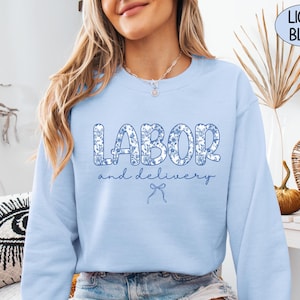 Op de afbeelding: Lichtblauw sweatshirt met de tekst "LABOR and delivery" in een blauwe bloemenprint. Het sweatshirt heeft een ronde hals en lange mouwen. Een lichtblauwe strik-afbeelding staat onder de tekst. De woorden "LIGHT BLUE" staan in een lichtblauwe cirkel.