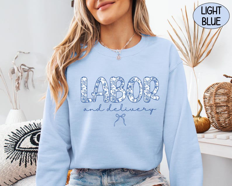 Op de afbeelding: Lichtblauw sweatshirt met de tekst "LABOR and delivery" in een blauwe bloemenprint. Het sweatshirt heeft een ronde hals en lange mouwen. Een lichtblauwe strik-afbeelding staat onder de tekst. De woorden "LIGHT BLUE" staan in een lichtblauwe cirkel.