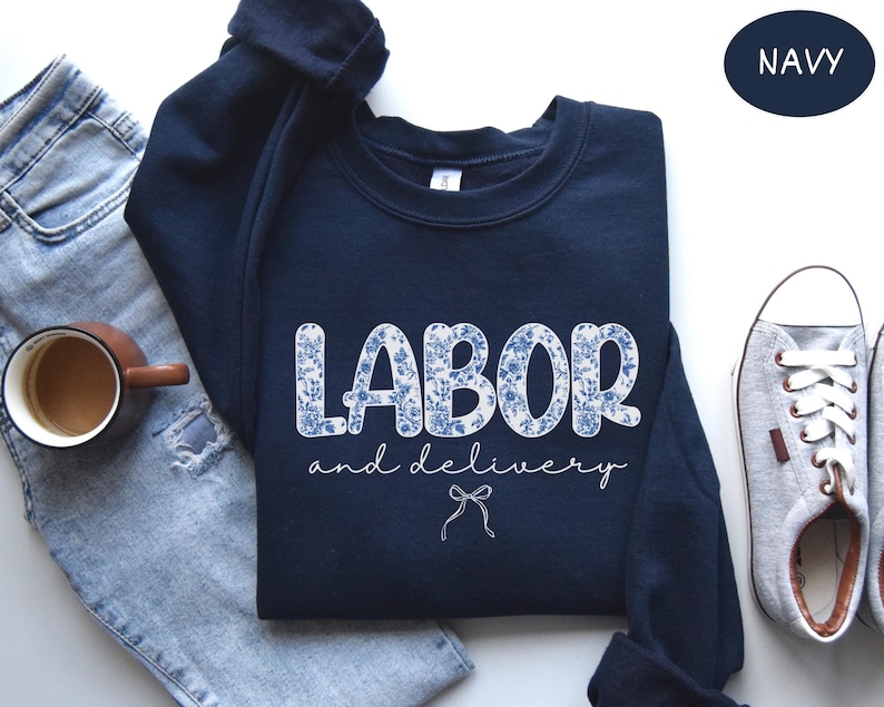 Op de afbeelding: Marineblauw sweatshirt met de tekst "LABOR and delivery" in witte, bloemmotief letters. Een kleine strik-afbeelding staat onder de tekst. Een kopje koffie, jeans en sneakers staan op de achtergrond.