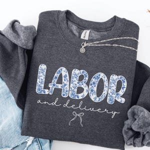 Op de afbeelding: Een donkergrijze sweatshirt met de woorden "LABOR" en "and delivery" in witte letters. Het woord "LABOR" is gevuld met een blauw bloemenpatroon. Een zilveren ketting met een kleine hanger ligt op de sweatshirt.