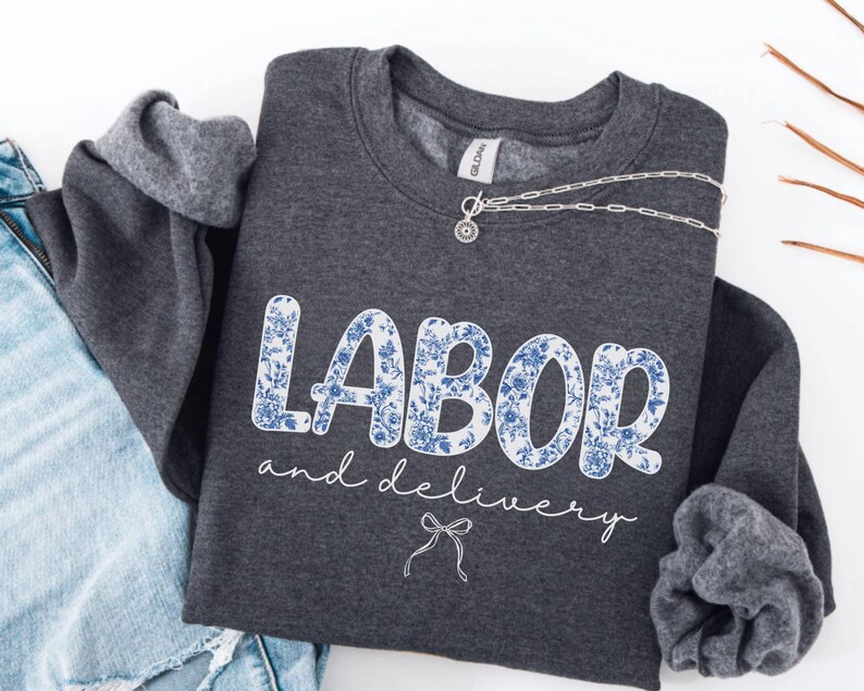 Op de afbeelding: Een donkergrijze sweatshirt met de woorden "LABOR" en "and delivery" in witte letters. Het woord "LABOR" is gevuld met een blauw bloemenpatroon. Een zilveren ketting met een kleine hanger ligt op de sweatshirt.