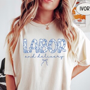 Op de afbeelding: Ivoor kleurig T-shirt met de tekst "LABOR and delivery" in blauwe bloemenprint. De tekst staat in het midden, met een kleine strik eronder. Het shirt heeft een losse pasvorm.
