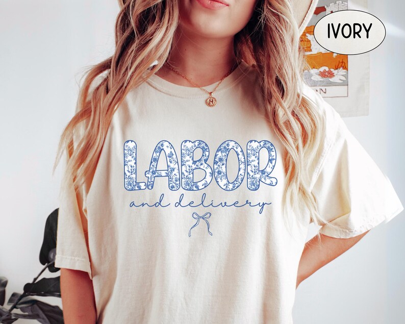 Op de afbeelding: Ivoor kleurig T-shirt met de tekst "LABOR and delivery" in blauwe bloemenprint. De tekst staat in het midden, met een kleine strik eronder. Het shirt heeft een losse pasvorm.