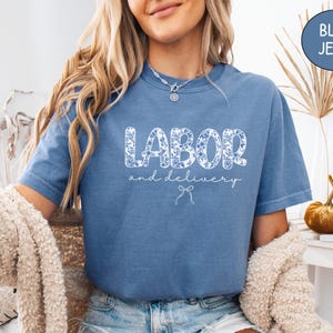 Op de afbeelding: Een blauw spijkerbroekkleurig t-shirt met de woorden "LABOR and delivery" in een bloemenpatroon. Het shirt heeft korte mouwen en een relaxte pasvorm. Een kleine strik-afbeelding staat onder de tekst.