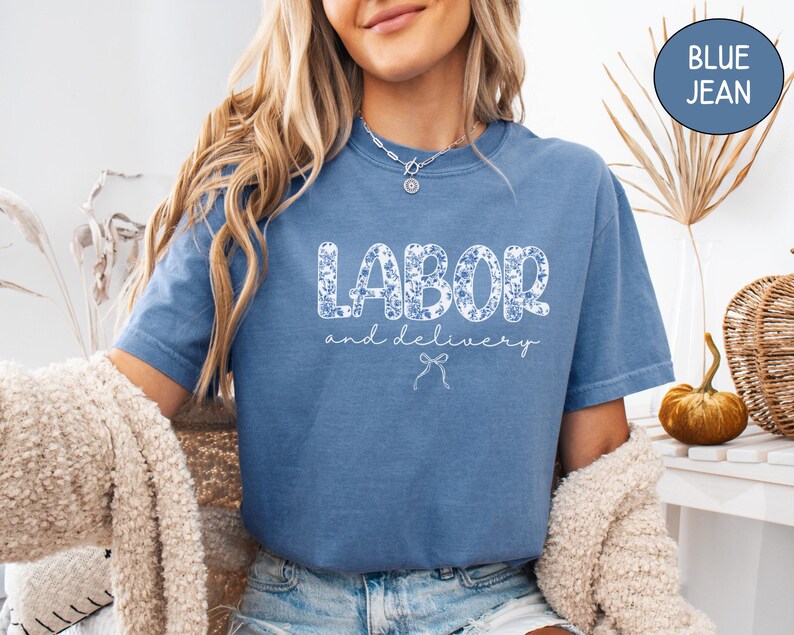 Op de afbeelding: Een blauw spijkerbroekkleurig t-shirt met de woorden "LABOR and delivery" in een bloemenpatroon. Het shirt heeft korte mouwen en een relaxte pasvorm. Een kleine strik-afbeelding staat onder de tekst.