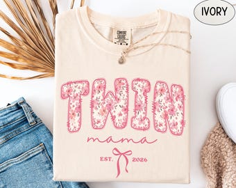 May include: An ivory t-shirt featuring the word TWIN in large block letters filled with a pink floral pattern. Below, the word mama is written in cursive, followed by a pink bow graphic and the text EST. 2026. The shirt has a Comfort Colors label.