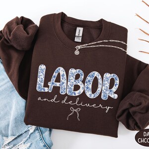 Op de afbeelding: Een donkerchocoladekleurige sweatshirt met de woorden "LABOR and delivery" in een blauw bloemenpatroon. Een zilveren ketting met een hanger is over de sweatshirt gedrapeerd. De sweatshirt is naast een spijkerbroek gevouwen.