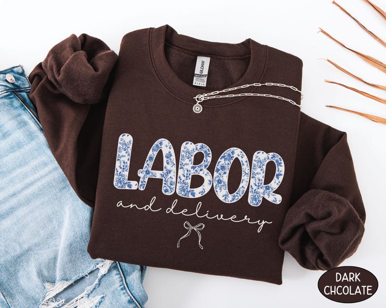 Op de afbeelding: Een donkerchocoladekleurige sweatshirt met de woorden "LABOR and delivery" in een blauw bloemenpatroon. Een zilveren ketting met een hanger is over de sweatshirt gedrapeerd. De sweatshirt is naast een spijkerbroek gevouwen.