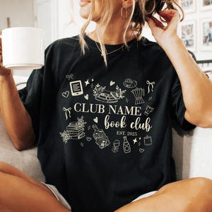 May include: Black t-shirt with a white graphic design featuring the text "CLUB NAME book club EST. 2025" and various book-related illustrations. The person is holding a white mug.