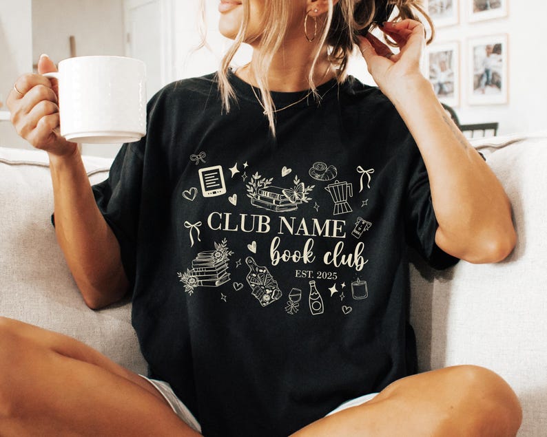 May include: Black t-shirt with a white graphic design featuring the text "CLUB NAME book club EST. 2025" and various book-related illustrations. The person is holding a white mug.