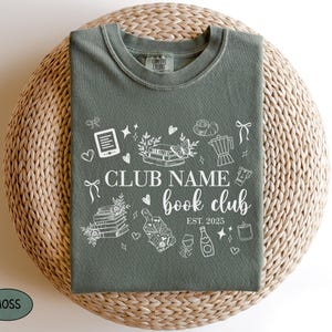 May include: A moss green t-shirt with a white graphic design featuring the words "Club Name book club" and the year "EST. 2025." The design includes illustrations of books, hearts, and other book-related items.
