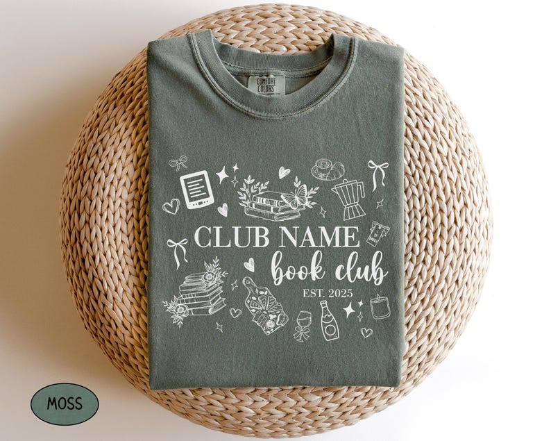 May include: A moss green t-shirt with a white graphic design featuring the words "Club Name book club" and the year "EST. 2025." The design includes illustrations of books, hearts, and other book-related items.