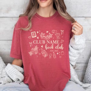 May include: A red-orange t-shirt with a white graphic design for a book club. The design includes the text "CLUB NAME book club EST 2025" with illustrations of books, hearts, and other related items.