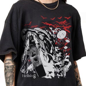 May include: Dark gray t-shirt featuring a black and white graphic of a character with a bottle, bats, and the text "Hellsing" in white. Red bats are in the background. The shirt has short sleeves.