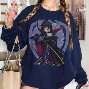 May include: Navy blue sweatshirt featuring a graphic of an anime character in a black and red cape. The character is set against a purple and white circular emblem. The sweatshirt has long sleeves.