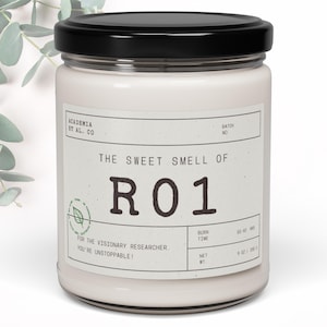May include: A white candle with a black lid and a label that reads "ACADEMIA ET AL. CO" and "THE SWEET SMELL OF R01". The label also includes the text "FOR THE VISIONARY RESEARCHER. YOU'RE UNSTOPPABLE!" and "BURN TIME 50-60 HRS NET WT 9 OZ | 255 G".